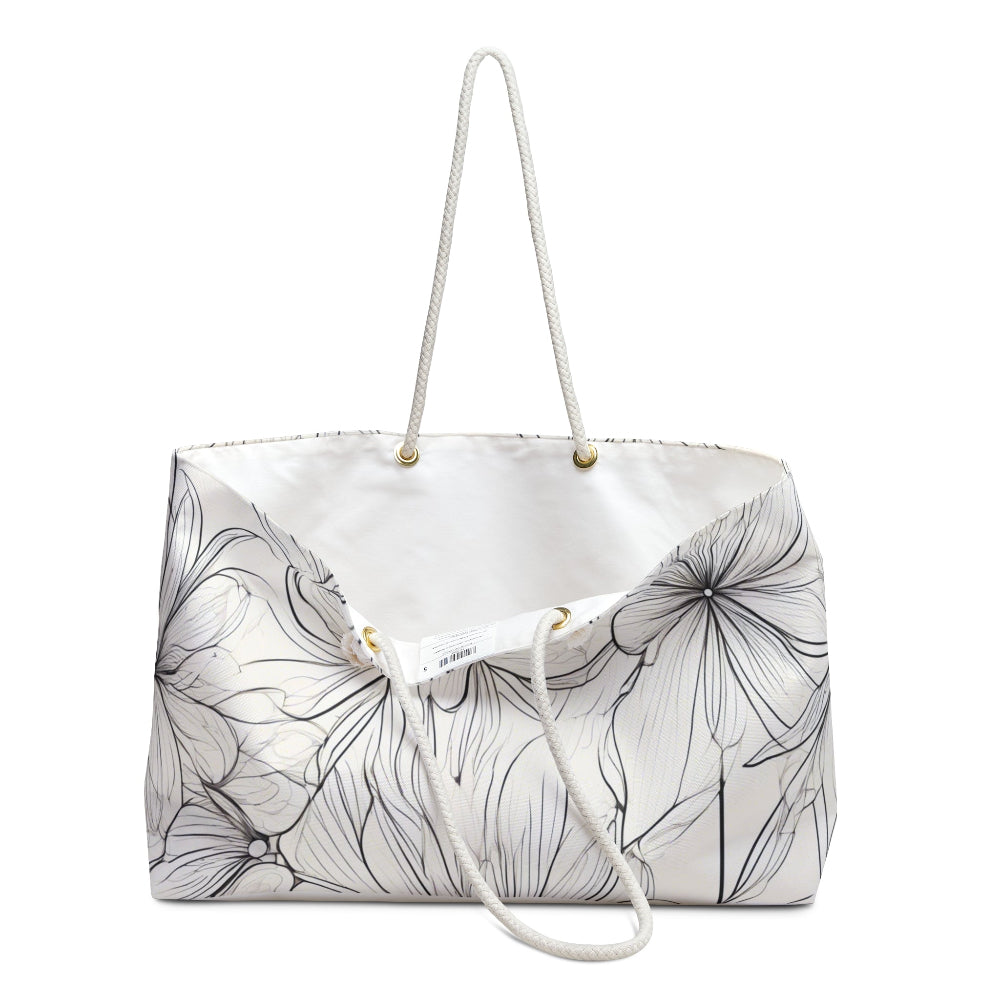 A white tote bag featuring an intricate black floral line drawing with delicate outlined flowers, equipped with white rope handles attached to gold eyelets—perfect for stylish everyday use